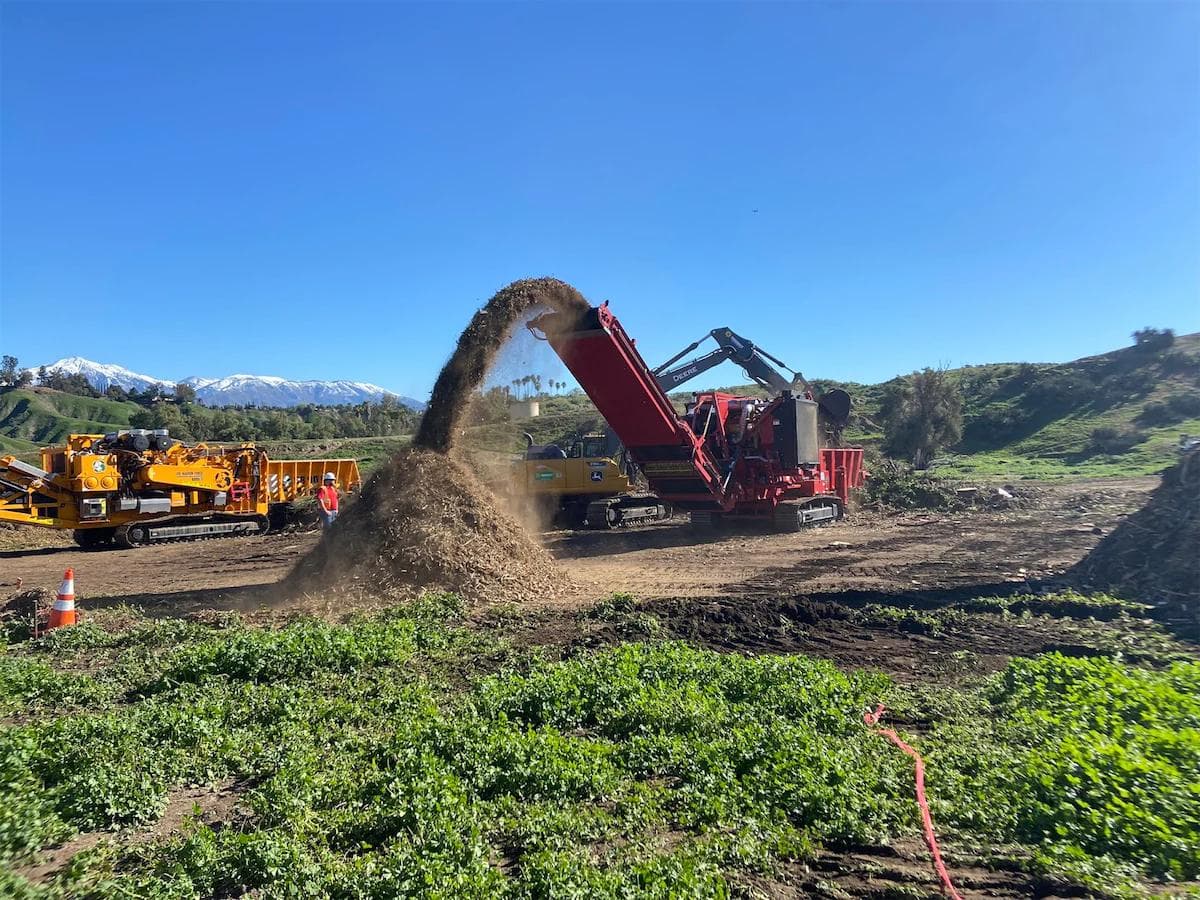 large red horizontal grinder shredding wood wase into a huge pile. the grinder is being fed by a yellow john deer excavator